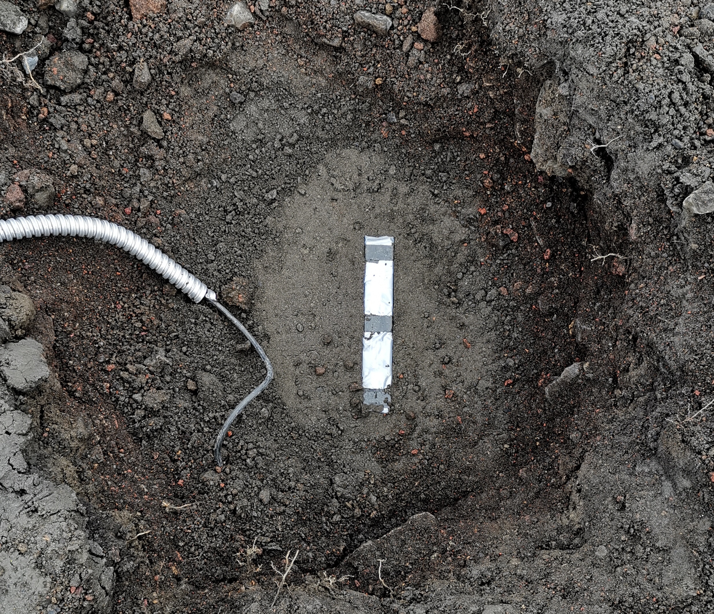 A top-down picture of a magnetometer in the ground. This is a rectangular box with dimensions of around 3 by 3 by 25 cm, surrounded by grainy, ashy, dark grey earth. The power and data cable is seen on the left-hand side going into a protective conduit.